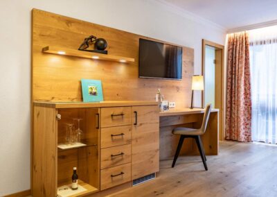Arbeitsbereich mit Holzkommode, Wand-TV, Schreibtisch, Stuhl und Regal mit Dekoration und Getränkeflasche.