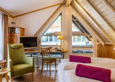 Dachgeschosszimmer mit großem Dreiecksfenster, Doppelbett mit pinken Deckenrollen, grünem Sessel, Schreibtisch und Fernseher.