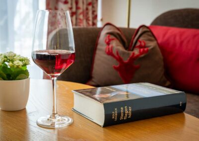 Detailaufnahme: Glas Rotwein, Buch, Kissen mit Hirschmotiv auf dem Sofa.
